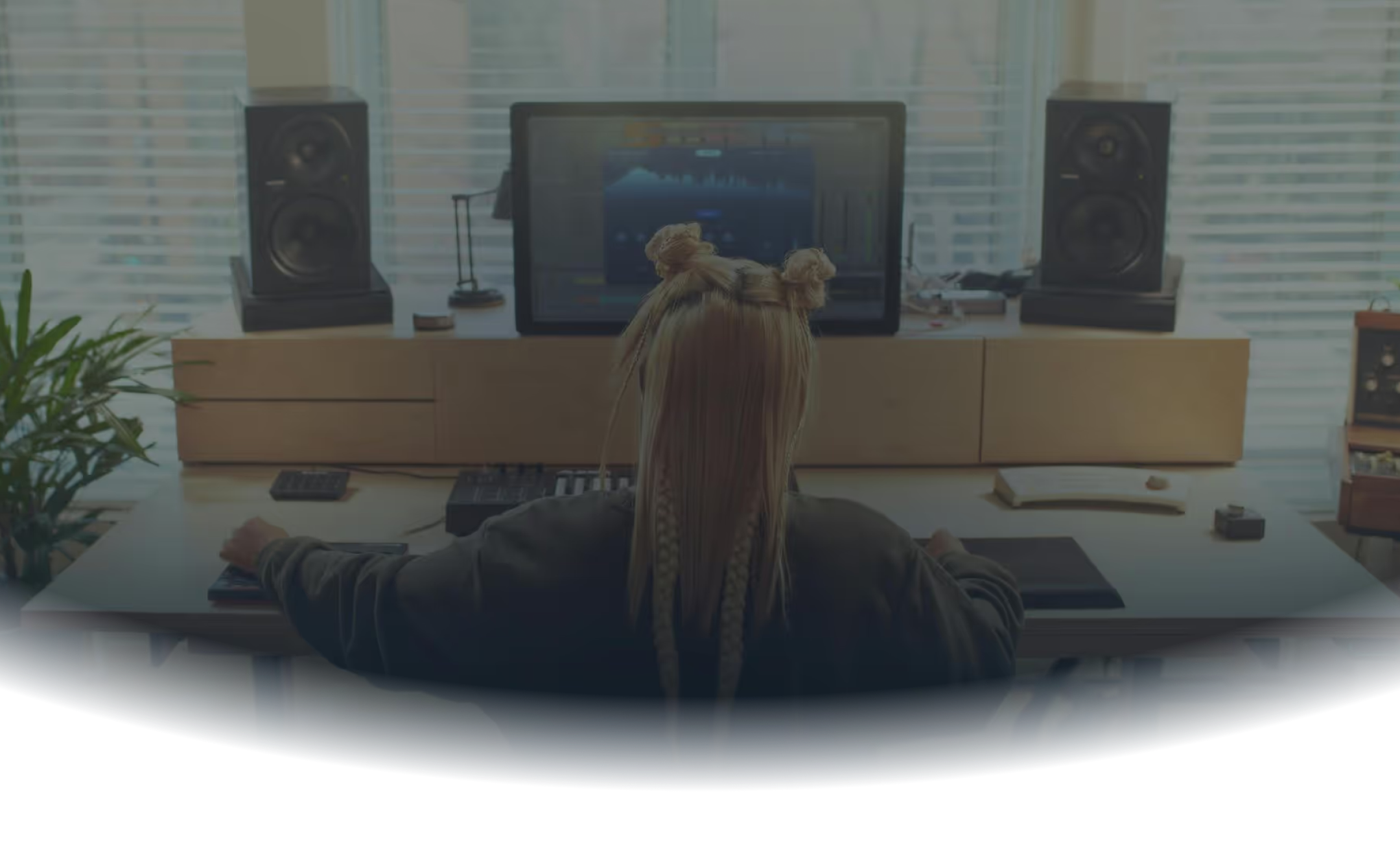 Person with braided hair styled in two buns working at a music production setup with speakers and a computer screen displaying audio software.